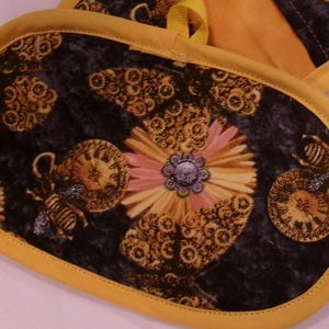 K&ouml;nnte beinhalten: Ovale Ofenhandschuhe mit gelbem Rand und Schlaufe. Die Handschuhe haben einen dunklen Hintergrund mit einem Blumen- und Bienenmuster in Gold-, Rosa- und Blaut&ouml;nen. Das Design umfasst Zifferbl&auml;tter und Zahnr&auml;der.