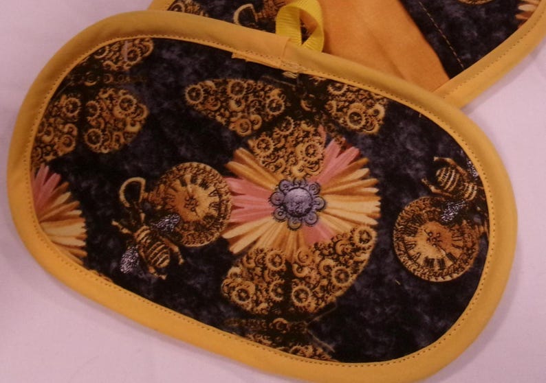 K&ouml;nnte beinhalten: Ovale Ofenhandschuhe mit gelbem Rand und Schlaufe. Die Handschuhe haben einen dunklen Hintergrund mit einem Blumen- und Bienenmuster in Gold-, Rosa- und Blaut&ouml;nen. Das Design umfasst Zifferbl&auml;tter und Zahnr&auml;der.