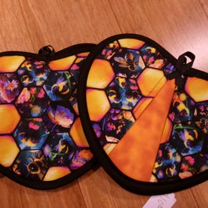 May include: Two heart-shaped pot holders with a black trim. The pot holders feature a honeycomb pattern with yellow and orange hexagons. The hexagons contain images of bees, flowers, and a dark background.
