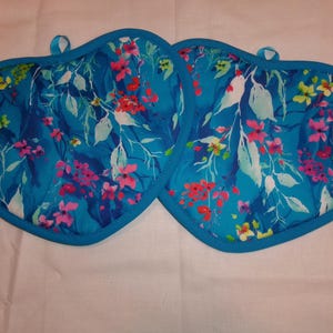 May include: Two heart-shaped pot holders with a vibrant floral pattern. The design features pink, red, and yellow flowers with white leaves on a teal background. Each pot holder has a teal border and a small loop for hanging.