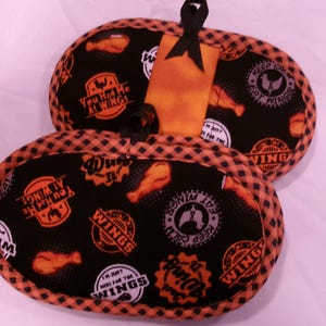 K&ouml;nnte beinhalten: Schwarze und orangefarbene Ofenhandschuhe mit H&uuml;hnerfl&uuml;gel-Motiv. Die Handschuhe haben einen schwarzen Hintergrund mit orangefarbenem und wei&szlig;em Text und H&uuml;hnerfl&uuml;gel-Grafiken. Der Text enth&auml;lt S&auml;tze wie "You Had Me At Wings" und "Keep Calm Eat Wings".