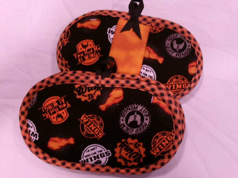 K&ouml;nnte beinhalten: Schwarze und orangefarbene Ofenhandschuhe mit H&uuml;hnerfl&uuml;gel-Motiv. Die Handschuhe haben einen schwarzen Hintergrund mit orangefarbenem und wei&szlig;em Text und H&uuml;hnerfl&uuml;gel-Grafiken. Der Text enth&auml;lt S&auml;tze wie "You Had Me At Wings" und "Keep Calm Eat Wings".