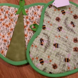 May include: Two heart-shaped pot holders. One features a floral pattern with orange flowers, the other has a green gingham pattern with bees and pinecones. Both have green trim and hanging loops.