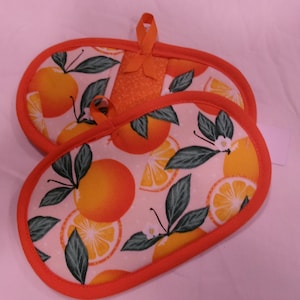 May include: Two orange and white oval-shaped pot holders with orange trim and hanging loops. The fabric features a pattern of oranges, orange slices, green leaves, and white blossoms. The pot holders are designed for kitchen use.