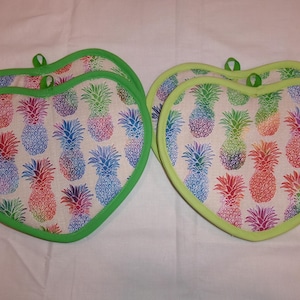 May include: Heart-shaped pot holders with a repeating pattern of colorful pineapples on a cream background. The edges are trimmed with a bright green fabric and have a loop for hanging.
