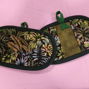 May include: Two oval pot holders with a tropical leaf design. They have a dark green border and a green loop for hanging. One pot holder has a solid green panel. The fabric features green, yellow, and blue leaves.