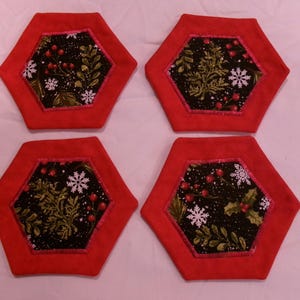May include: Set of four hexagonal coasters. Each coaster has a red border and a black center with a festive design of green leaves, red berries, and white snowflakes. The coasters are made of fabric.