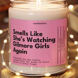 May include: A scented soy wax candle in a clear glass jar with a pink label. The label reads: sweetwickco, Smells Like She’s Watching Gilmore Girls Again, A Scented Soy Wax Candle, Infused With The Aroma Of Stars, Hollow In Every Flame, Soy Wax Blend.
