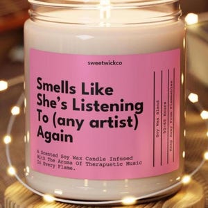 Puede incluir: Una vela en frasco de vidrio transparente con cera de soja color blanco hueso. La etiqueta rosa tiene texto negro que dice: Smells Like She's Listening To (any artist) Again, sweetwickco, Soy Wax Blend, 50-60 Hours, Keep Away From Flammables, A Scented Soy Wax Candle Infused With The Aroma Of Therapuetic Music In Every Flame.