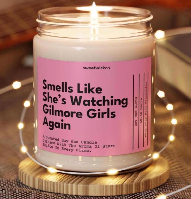 May include: A scented soy wax candle in a clear glass jar with a pink label. The label reads: sweetwickco, Smells Like She’s Watching Gilmore Girls Again, A Scented Soy Wax Candle, Infused With The Aroma Of Stars, Hollow In Every Flame, Soy Wax Blend.