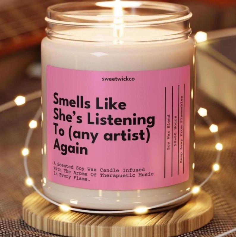 Puede incluir: Una vela en frasco de vidrio transparente con cera de soja color blanco hueso. La etiqueta rosa tiene texto negro que dice: Smells Like She's Listening To (any artist) Again, sweetwickco, Soy Wax Blend, 50-60 Hours, Keep Away From Flammables, A Scented Soy Wax Candle Infused With The Aroma Of Therapuetic Music In Every Flame.