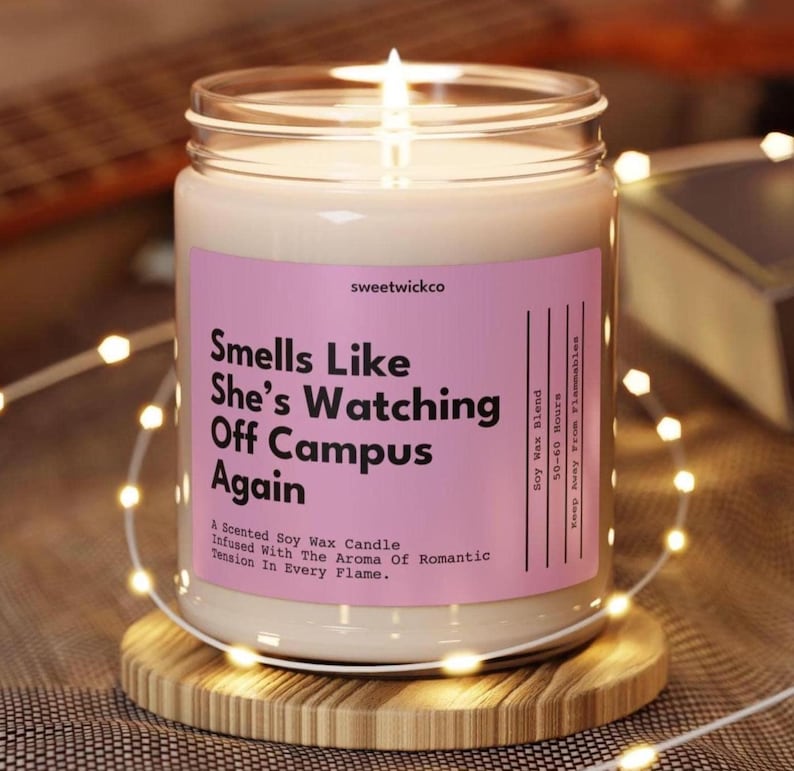 May include: A clear glass jar candle with a light-colored soy wax blend and a lit wick. The pink label features the text: "sweetwickco", "Smells Like She's Watching Off Campus Again", "A Scented Soy Wax Candle", "Infused With The Aroma Of Romantic Tension In Every Flame", "Soy Wax Blend", and "50-60 Hours".