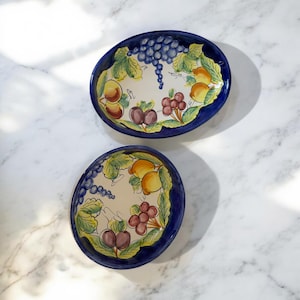 May include: Two ceramic bowls with blue rims and a white interior. The bowls are decorated with a colorful fruit design featuring grapes, plums, lemons, and leaves.