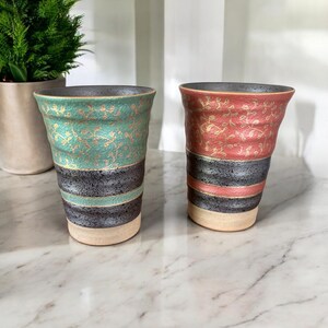 May include: Two ceramic cups with a black and gold striped design. One cup has a teal floral pattern and the other has a pink floral pattern.