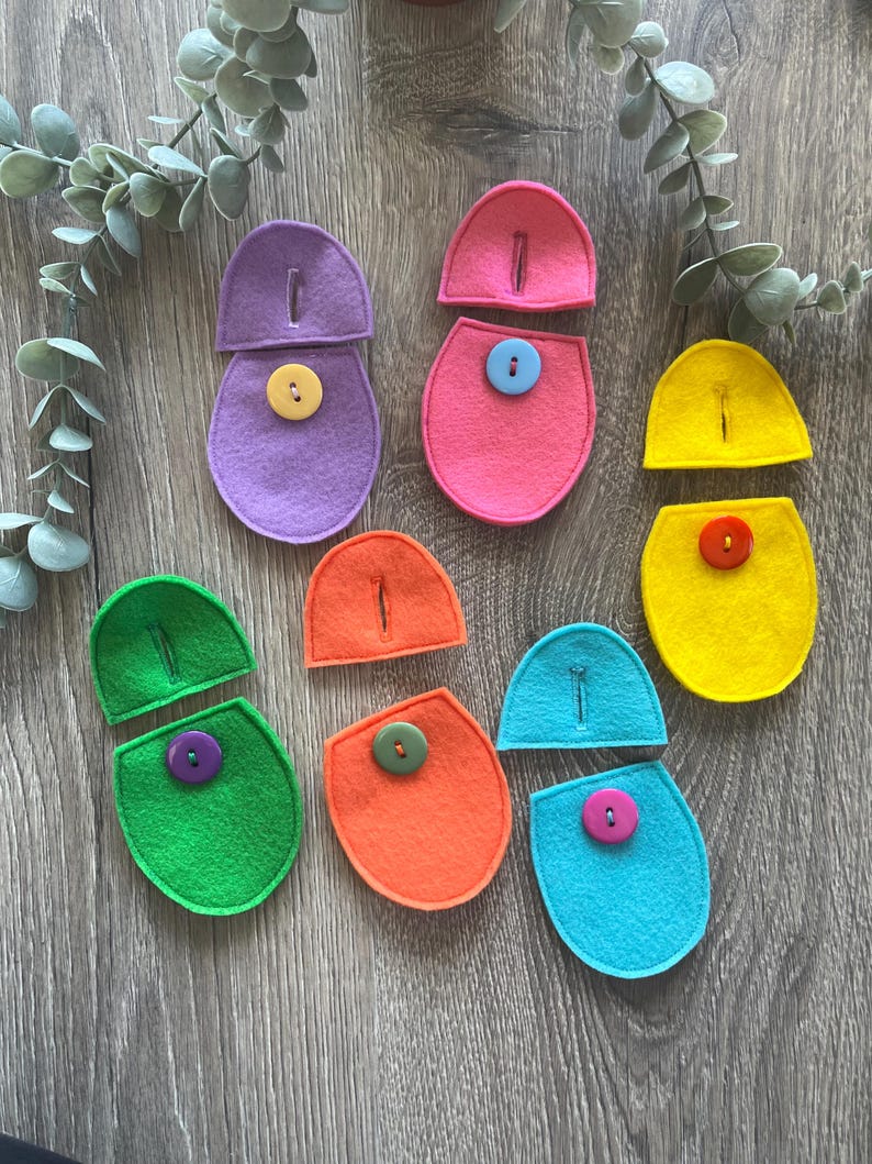 Op de afbeelding: Zes viltvormen met knopen in verschillende kleuren. Elke vorm is in twee&euml;n gesplitst, met een helft met een knoop en de andere helft met een gleuf voor de knoop. De kleuren zijn paars, roze, geel, groen, oranje en blauw.