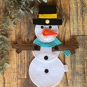 May include: A felt snowman craft with a black top hat, a yellow band, white face, orange nose, blue scarf, brown twig arms, and black buttons. The snowman is on a wood background.