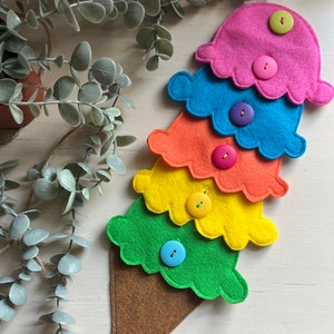 May include: A felt ice cream cone toy with five scoops of ice cream in rainbow colors. Each scoop has a button in the center. The cone is brown and the scoops are pink, blue, orange, yellow, and green.