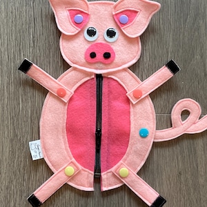 May include: A light pink felt pig toy featuring a central black zipper, googly eyes, and a dark pink snout. The pig has four limbs attached with colorful plastic snaps, black felt hooves, a curly tail, and a small white tag labeled Sweet Peach OT.