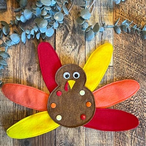 May include: A felt craft turkey with a brown body, red, orange, and yellow feathers, and googly eyes. The turkey has a red beak and brown buttons for details.