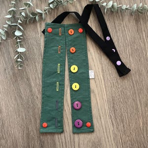 May include: Green fabric suspenders with colorful buttons in orange, yellow, and purple. The suspenders have a black strap and are decorated with embroidered rectangles. The suspenders are laying on a wooden surface.
