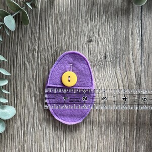 Op de afbeelding: Paarse vilten ei met een gele knoop. Het ei is ongeveer 5 cm lang en wordt gemeten met een liniaal.