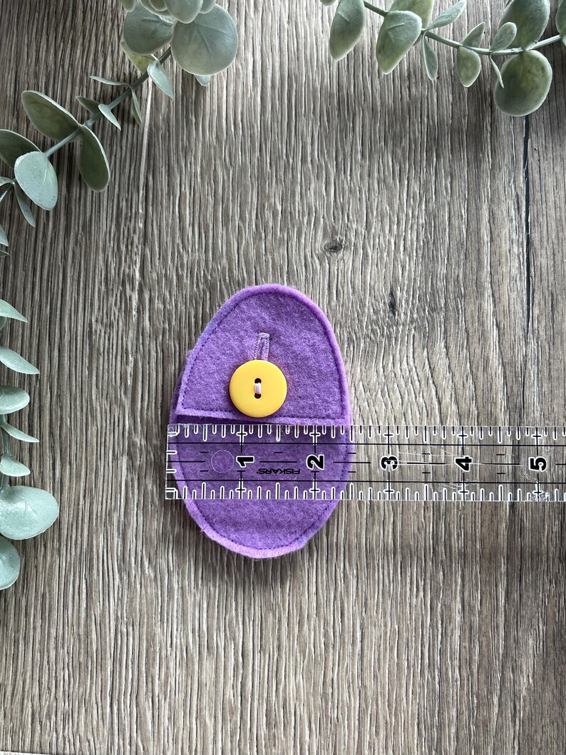 Op de afbeelding: Paarse vilten ei met een gele knoop. Het ei is ongeveer 5 cm lang en wordt gemeten met een liniaal.