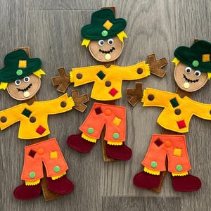 May include: Three felt scarecrow decorations featuring dark green hats, tan circular faces with googly eyes, yellow shirts with colorful patches, and orange pants with green accents. Each figure is mounted on a brown vertical wooden stake.