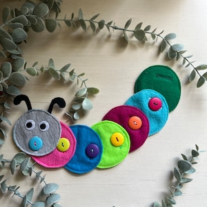 May include: A colorful felt caterpillar toy with buttons for eyes and a row of colorful buttons down its body. The caterpillar is made of felt and has a gray head with googly eyes.