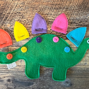 May include: A green felt dinosaur toy with colorful button accents and removable felt plates. The dinosaur has a button eye and a small tag. The removable plates are orange, yellow, purple, pink, and blue.