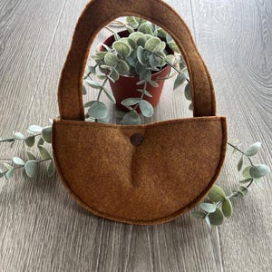 Op de afbeelding: Een bruine wollen wandhanger met een afgeronde bodem en een lusvormige handgreep. De hanger is leeg en klaar om een kleine plant te houden.