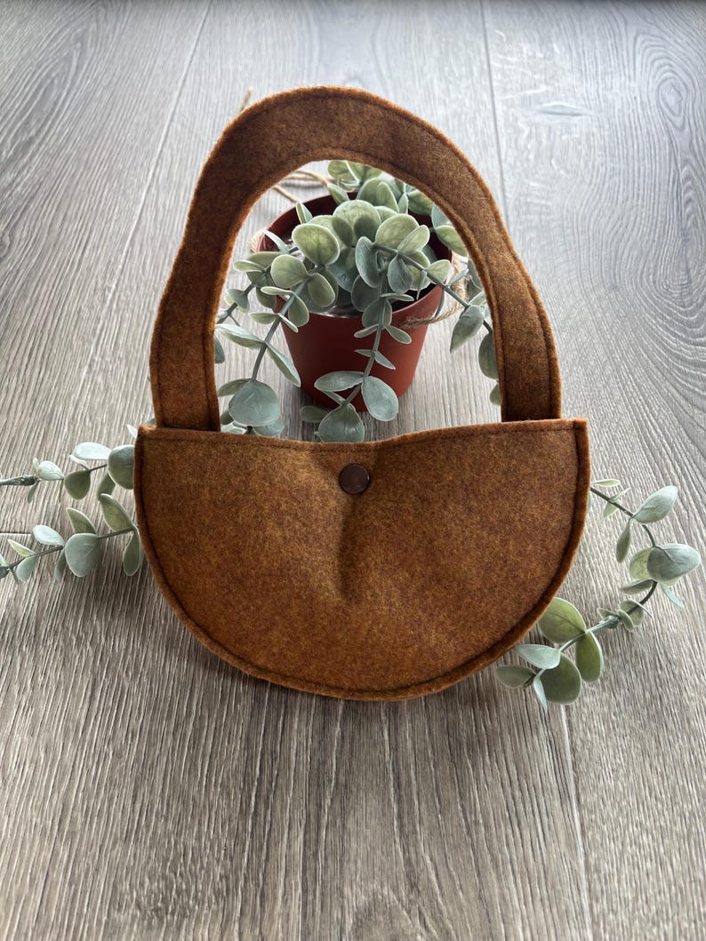 Op de afbeelding: Een bruine wollen wandhanger met een afgeronde bodem en een lusvormige handgreep. De hanger is leeg en klaar om een kleine plant te houden.