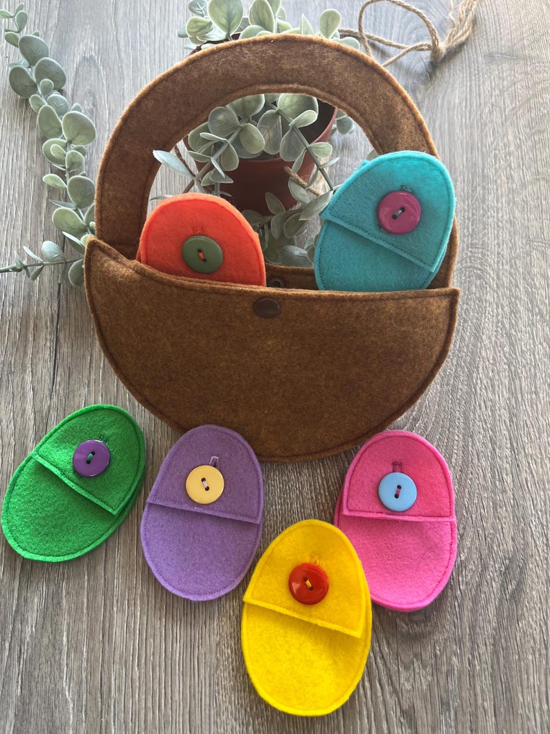 Op de afbeelding: Een bruine vilten tas met een handvat, met zes vilten, eivormige zakjes met verschillende kleuren vilt en knopen. De zakjes zijn in de kleuren groen, paars, roze, geel, oranje en blauw.