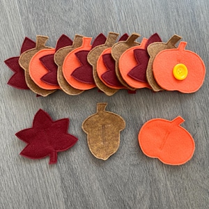May include: A collection of felt shapes including maroon maple leaves, brown acorns, and orange pumpkins. Some pieces feature vertical slits for attachment, and one orange pumpkin is adorned with a single, bright yellow four-hole button.
