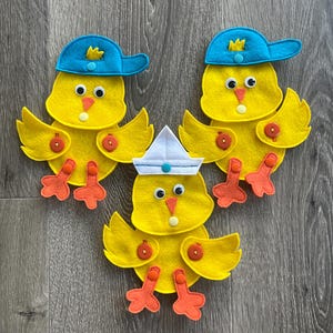 May include: Three yellow felt chick decorations with orange feet and beaks. Two chicks wear blue baseball caps with yellow crowns, while the third wears a white sailor hat. The chicks have button accents and are set against a wood grain background.