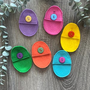 Op de afbeelding: Zes vilten eivormige zakjes met verschillende kleuren vilt en knopen. De zakjes zijn paars, roze, geel, oranje, groen en blauw.