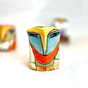 May include: A small ceramic figurine with a stylized face. The face is painted with yellow, blue, orange, and green colors. The figurine is a whimsical and colorful piece of art.