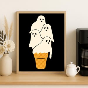 May include: A framed art print featuring a whimsical illustration of four white ghost figures stacked on top of a waffle cone against a black background. The artwork is in a wooden frame and is displayed on a wooden surface.