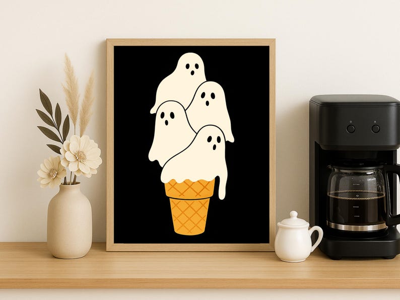 May include: A framed art print featuring a whimsical illustration of four white ghost figures stacked on top of a waffle cone against a black background. The artwork is in a wooden frame and is displayed on a wooden surface.