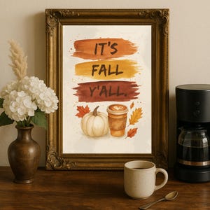 Może przedstawiać: Oprawiony wydruk artystyczny z napisem "IT'S FALL Y'ALL" w pomarańczowych, żółtych i brązowych pociągnięciach pędzla. Wydruk przedstawia akwarelową dynię, filiżankę kawy i jesienne liście. Na stole filiżanka do kawy, łyżeczka i ekspres do kawy.