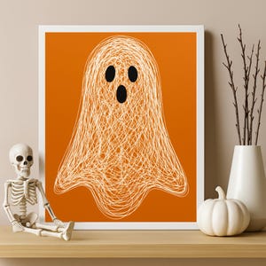 May include: A framed print with a white ghost illustration on an orange backdrop. The ghost is created with a scribbled design, featuring three black facial details. A white pumpkin, a skeleton figurine, and a vase with branches are in the foreground.