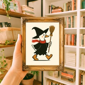 Op de afbeelding: Een ingelijste illustratie van een cartoon eend verkleed als heks, met een zwarte hoed, cape en laarzen, met een bezem. De eend heeft een rode sjaal en loopt. De lijst is goudkleurig.