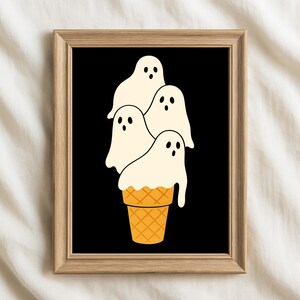 May include: A framed art print featuring a whimsical illustration of four ghost figures stacked on top of a waffle cone. The ghosts are white with black eyes and the cone is orange. The background is black.