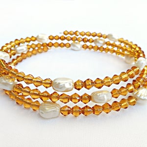 Puede incluir: Un brazalete de triple cadena hecho con cuentas de vidrio color ámbar y perlas blancas falsas. El brazalete está enrollado y listo para ser usado.