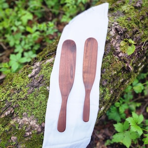 May include: Two wooden spatulas, one brown and one dark brown, are laid out on a white cloth. The cloth is on top of a mossy log.