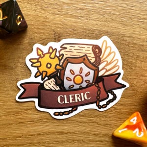 May include: A sticker featuring a cleric character from a tabletop role-playing game. The sticker includes a shield with a sun, a mace, a scroll, and a pair of wings. The sticker has a red banner with the word "CLERIC" written on it.