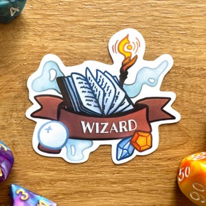 Könnte beinhalten: Ein weißer Aufkleber mit einem braunen Banner, auf dem "WIZARD" steht. Das Banner ist mit einem blauen, geöffneten Buch, einer weißen Kugel, einem blauen Kristall und einem orangefarbenen Kristall verziert. Aus dem Buch kommt eine Flamme.