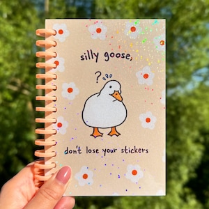 Könnte beinhalten: Ein Spiralnotizbuch mit pfirsichfarbener Bindung und Einband. Der Einband zeigt eine Cartoon-Gans-Illustration, den Text "silly goose, ?... don't lose your stickers", weiße Blumen und Glitzerakzente.
