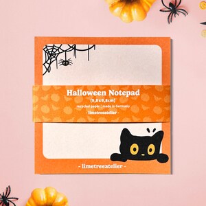 May include: Halloween notepad featuring a black cat illustration and spiderweb design. The notepad is orange and white, with the text "Halloween Notepad" and "(9.8x9.8cm)". Made from recycled paper.