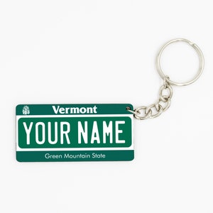 May include: Green and white keychain with a Vermont license plate design. The plate says "Vermont" at the top and "YOUR NAME" in large white letters. The bottom of the plate says "Green Mountain State".