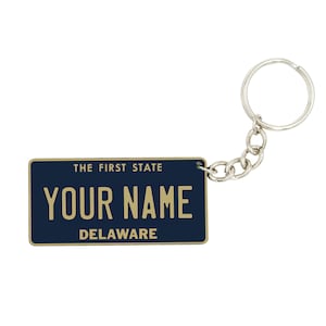 Peut inclure: Un porte-clés bleu et or en forme de plaque d'immatriculation du Delaware avec le texte "THE FIRST STATE" et "YOUR NAME" en lettres dorées. Le porte-clés a un anneau en métal argenté.